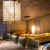 En stor, gul hjørnesofa er plassert rundt en glødende lampe i et svakt opplyst rom, med mønstrede tepper og komfortable stoler rundt et trebord. Stofflamper henger over.