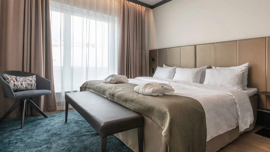 A neatly made bed with pillows and robes sits beside a window with beige curtains, surrounded by a chair, bench, and soft blue carpet in a cozy room.