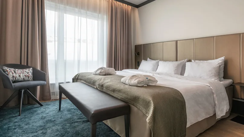 A neatly made bed with pillows and robes sits beside a window with beige curtains, surrounded by a chair, bench, and soft blue carpet in a cozy room.
