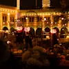Jack-o'-lantern-figurer står under ett upplyst paviljong, omgiven av pumpor och festliga ljus på natten. En upplyst byggnad reser sig i bakgrunden.