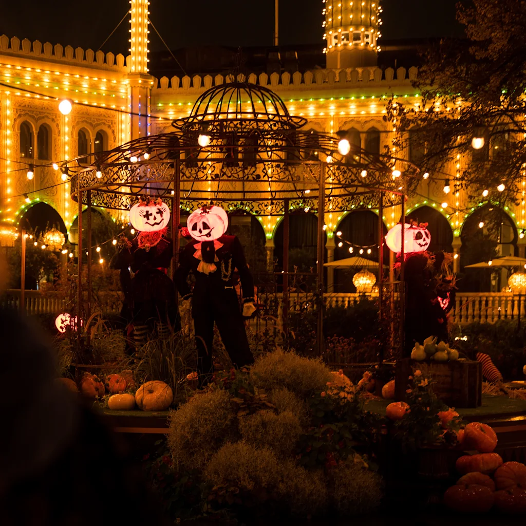 Jack-o'-lantern-figurer står under ett upplyst paviljong, omgiven av pumpor och festliga ljus på natten. En upplyst byggnad reser sig i bakgrunden.