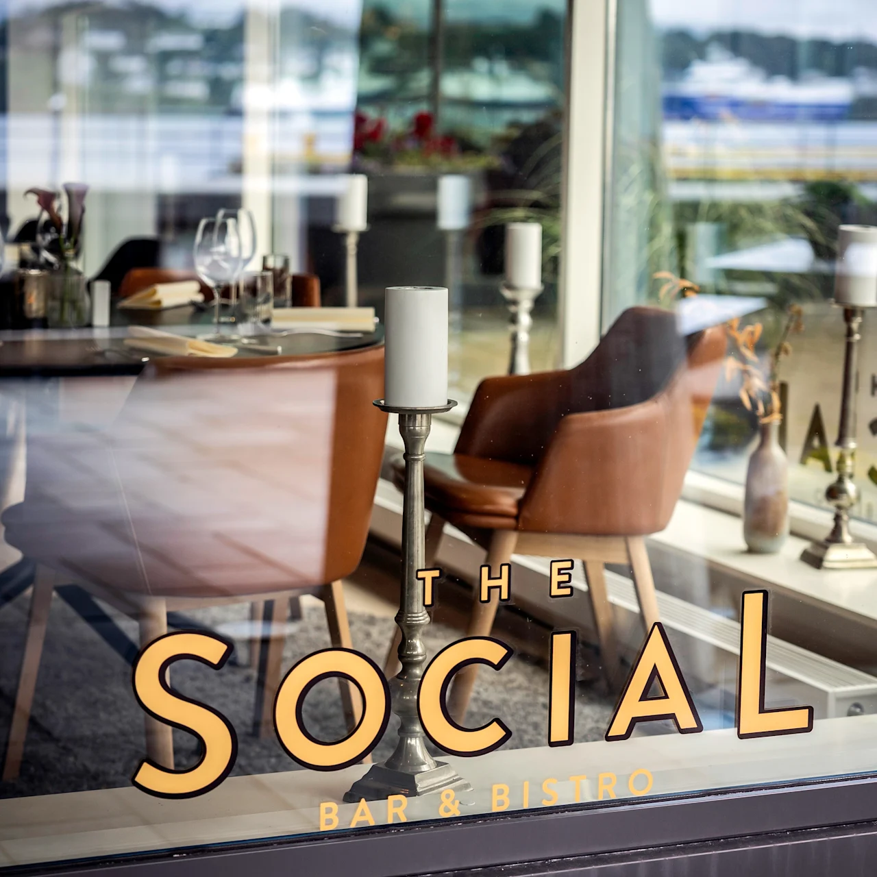 Bruna läderstolar runt bord med vinglas och bestick set genom ett fönster. "THE SOCIAL BAR & BISTRO" står på glaset med guldbokstäver.
