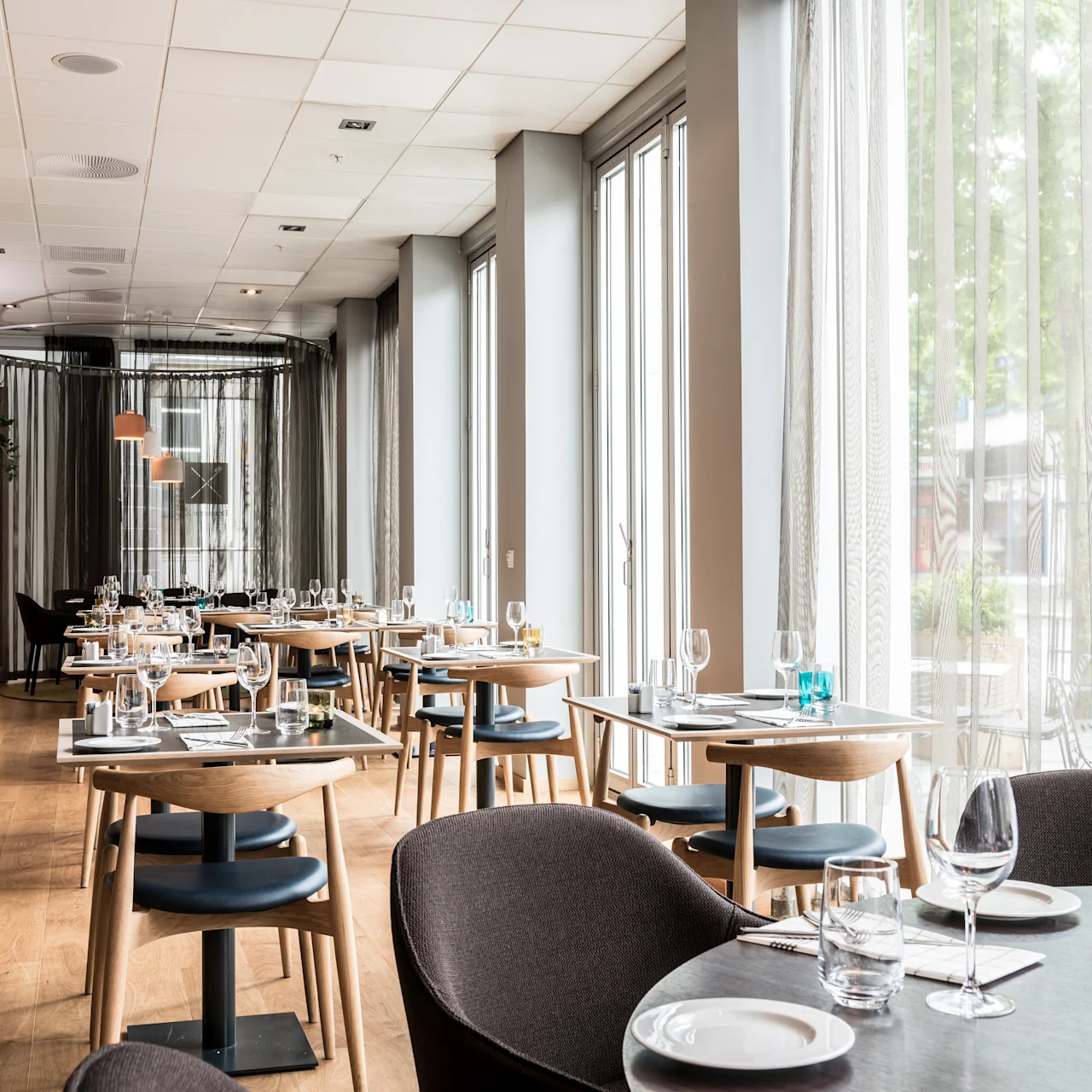 Moderni ravintola, jossa puiset tuolit ja pöydät on katettu ruokailua varten, ympäröity suurilla ikkunoilla ja valkoisilla verhoilla, luoden valoisan ilmapiirin.