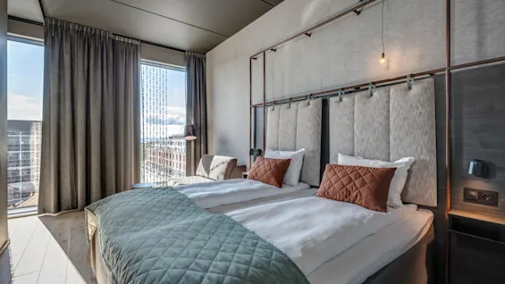 Ett modernt hotellrum med två prydligt arrangerade enkelsängar med gröna täcken och bruna kuddar, beläget under ett stort fönster med stadsutsikt och tunga gardiner.