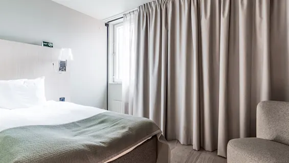 A bright and inviting hotel room with a comfortable bed, large window with curtains, and a cozy armchair.
