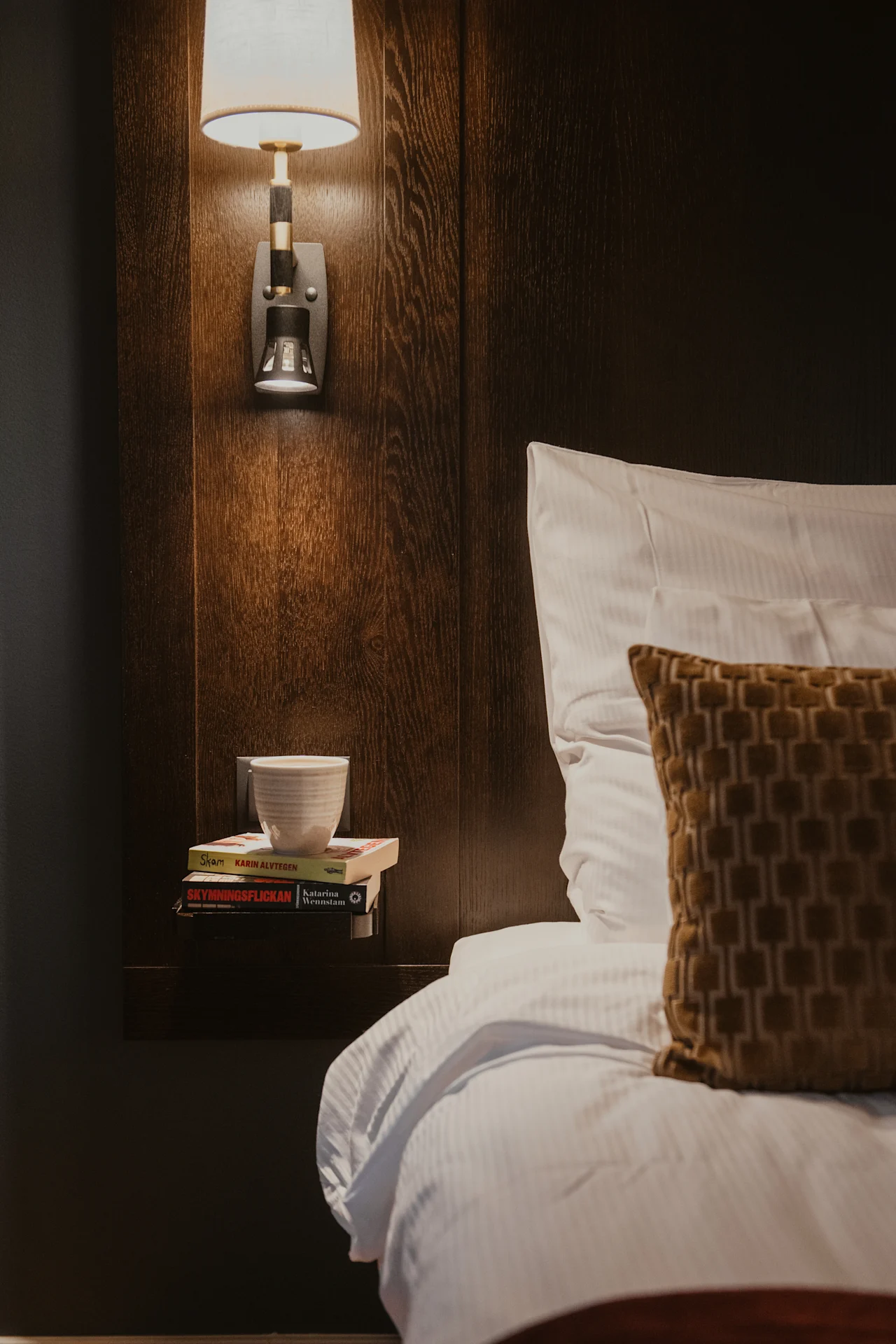 Cozy bedside at Home Hotel Kung Oscar with a warm lamp, books (SKAM, SKYMNINGSFLICKAN), and a cup, perfect for reading.