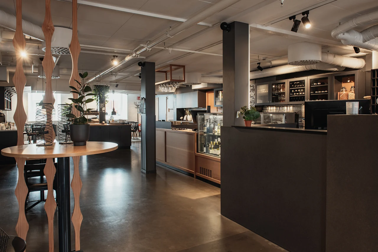 Moderni ja kutsuva hotelliravintola tai baarialue tummalla tiskillä, puupöydillä ja viherkasveilla avoimessa tilassa.