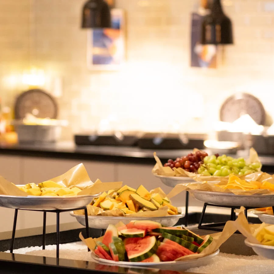 Plates of assorted fruits, including watermelon, grapes, and mango, arranged on a buffet table in a warmly lit dining area.