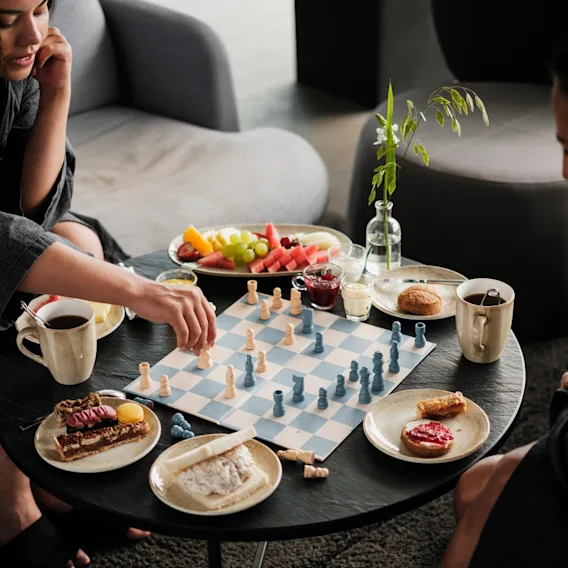Två personer spelar schack vid ett runt bord med olika frukostar, inklusive frukt och bakverk, och kaffemuggar, i en mysig inomhusmiljö.