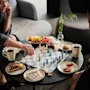 Två personer spelar schack vid ett runt bord med olika frukostar, inklusive frukt och bakverk, och kaffemuggar, i en mysig inomhusmiljö.