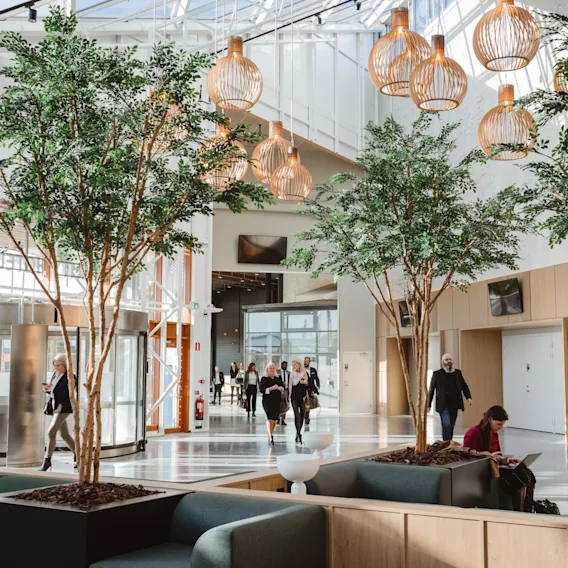 A bright, modern hotel lobby with people moving and relaxing. Indoor trees and unique lighting create a welcoming atmosphere. Arlanda.