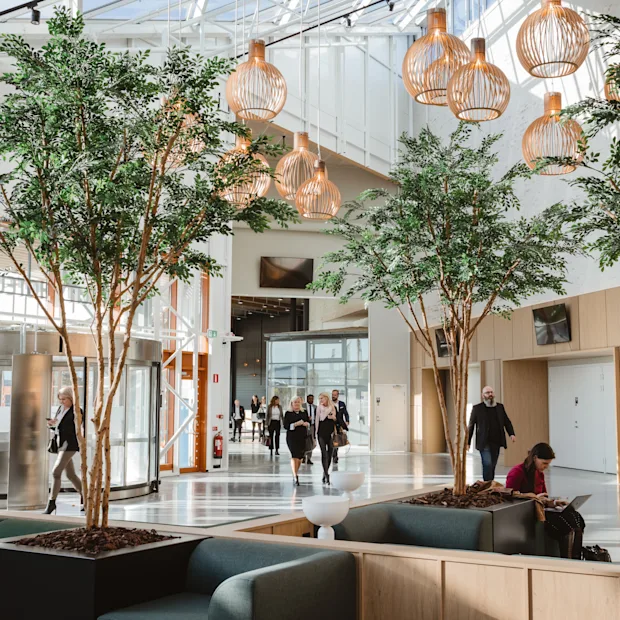 A bright, modern hotel lobby with people moving and relaxing. Indoor trees and unique lighting create a welcoming atmosphere. Arlanda.