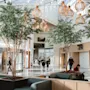 A bright, modern hotel lobby with people moving and relaxing. Indoor trees and unique lighting create a welcoming atmosphere. Arlanda.