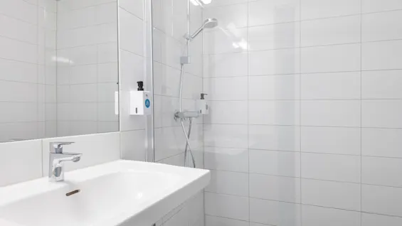 Handfat med kromkran som speglas i en badrumsspegel. Duschhuvud och tvåldispensrar monterade på kaklade väggar. Ljust belyst, minimalistisk vitkaklad miljö. Text på dispenser: "SHAMPOO". 