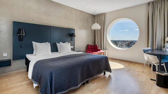 Et moderne hotelværelse med en stor seng, et rundt vindue med udsigt over byen og et lille arbejds- eller spiseområde.