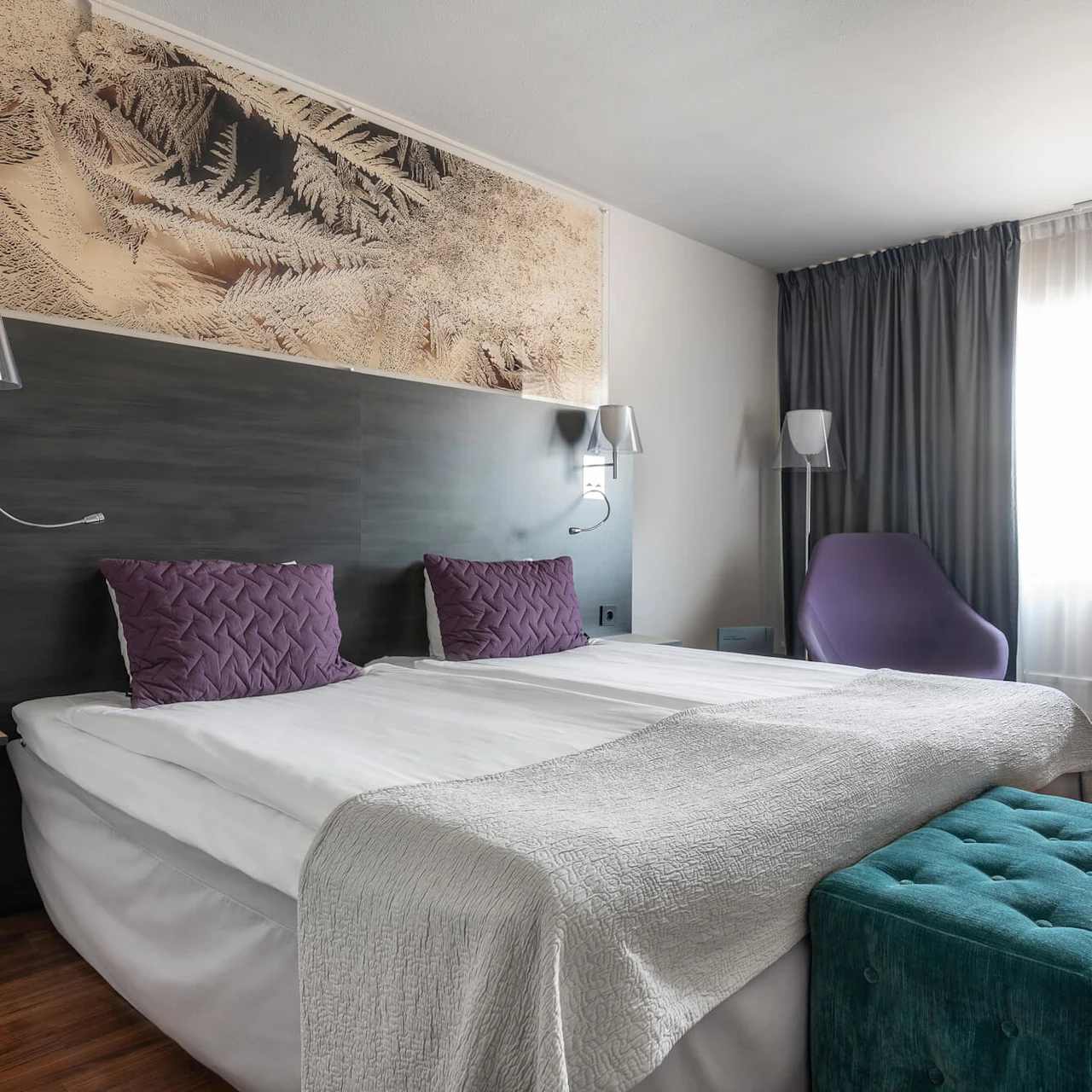 Ett modernt hotellrum har en stor säng med vita sängkläder och lila kuddar. Ett väggkonstverk och mjuk belysning framhäver den samtida inredningen, med ett fönster täckt av grå gardiner i närheten.