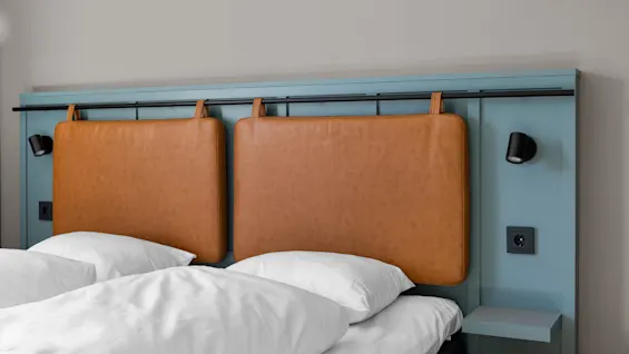 Sängyssä on sininen pääty ja kiinnitetyt ruskeat nahkatyynyt, joita reunustavat mustat lukuvalot. Valkoiset tyynyt ja vuodevaatteet täydentävät modernin ja minimalistisen makuuhuoneen.