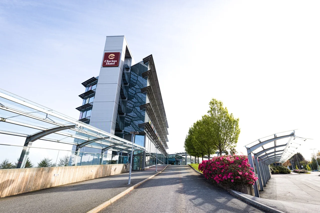 Et høyhus med et "Clarion Hotel"-skilt står i et moderne, beplantet område med grøntområder og blomster, ved siden av en glassgang under en klar himmel.