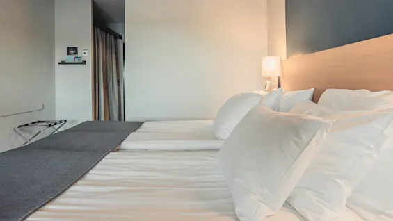 Clean white pillows neatly arranged on a double bed with a wooden headboard, in a softly-lit modern hotel room, featuring a small wall shelf and a luggage rack.
