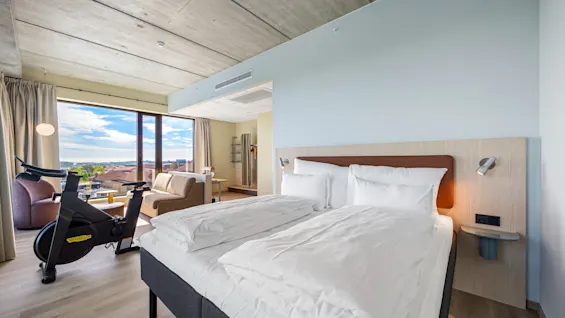 A modern hotel room with a bed, a sitting area, and an exercise bike by a large window with a scenic view.