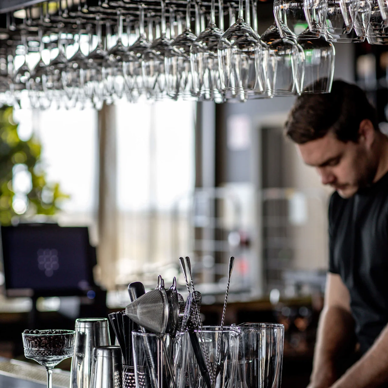 Glas hænger ovenfor, mens en bartender forbereder drinks i en bar. Forskellige redskaber, shakere og glas står på disken. Blød indendørs belysning skaber en reflekterende effekt på overfladen.