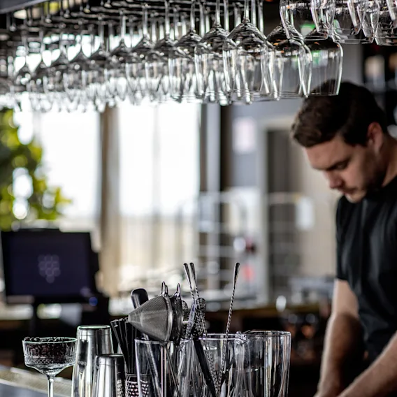 Glas hänger ovanför medan en bartender förbereder drycker vid en bar. Olika redskap, shakers och glas står på disken. Mjuk inomhusbelysning skapar en reflekterande effekt på ytan.