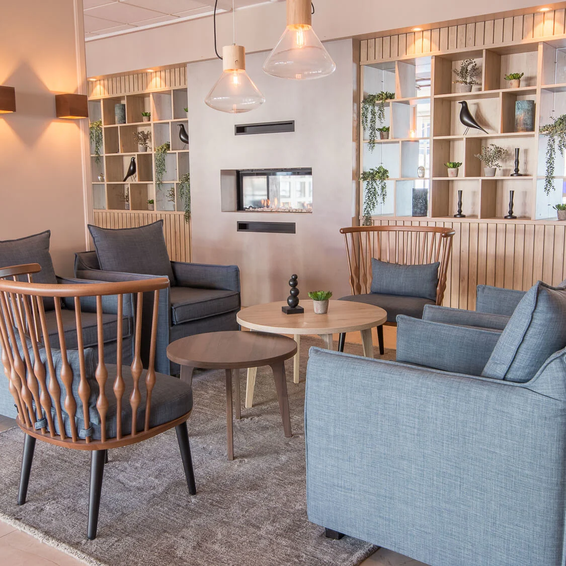 En koselig lounge med blå lenestoler rundt et trebord med hengelamper over. Moderne hyller med planter og dekorative gjenstander i bakgrunnen, som komplementerer den varme atmosfæren.
