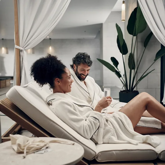 Wellness på Clarion Hotel. Kaksi kylpytakkiin pukeutunutta henkilöä rentoutuu ja hymyilee rauhallisessa kylpyläympäristössä.