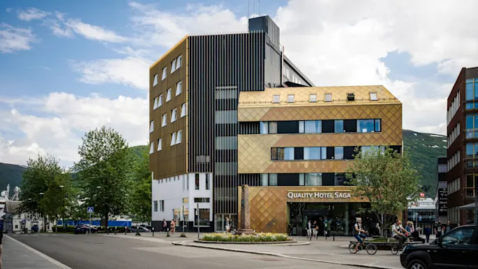 A building labeled "QUALITY HOTEL SAGA" stands in a bustling street context, featuring modern architecture with reflective panels and multiple windows; pedestrians and cyclists move nearby.