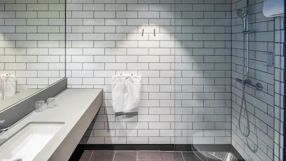 Et moderne bad har en vegg med hvite fliser, en glassdusj med kromdetaljer og en vask med to glass og speil. Håndklær henger pent på kroker.