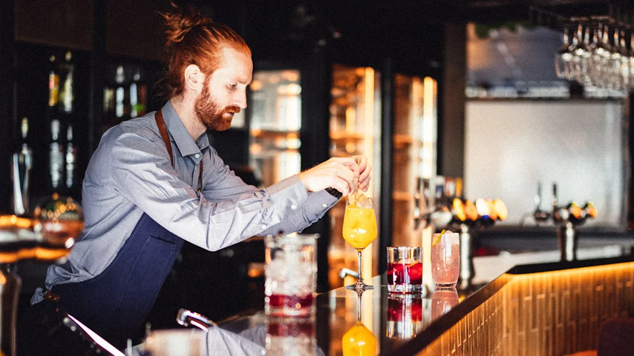 En bartender med oppsatt hår, iført blå skjorte og forkle, lager en cocktail i en svakt opplyst bar. Glass og flasker er synlige i bakgrunnen.