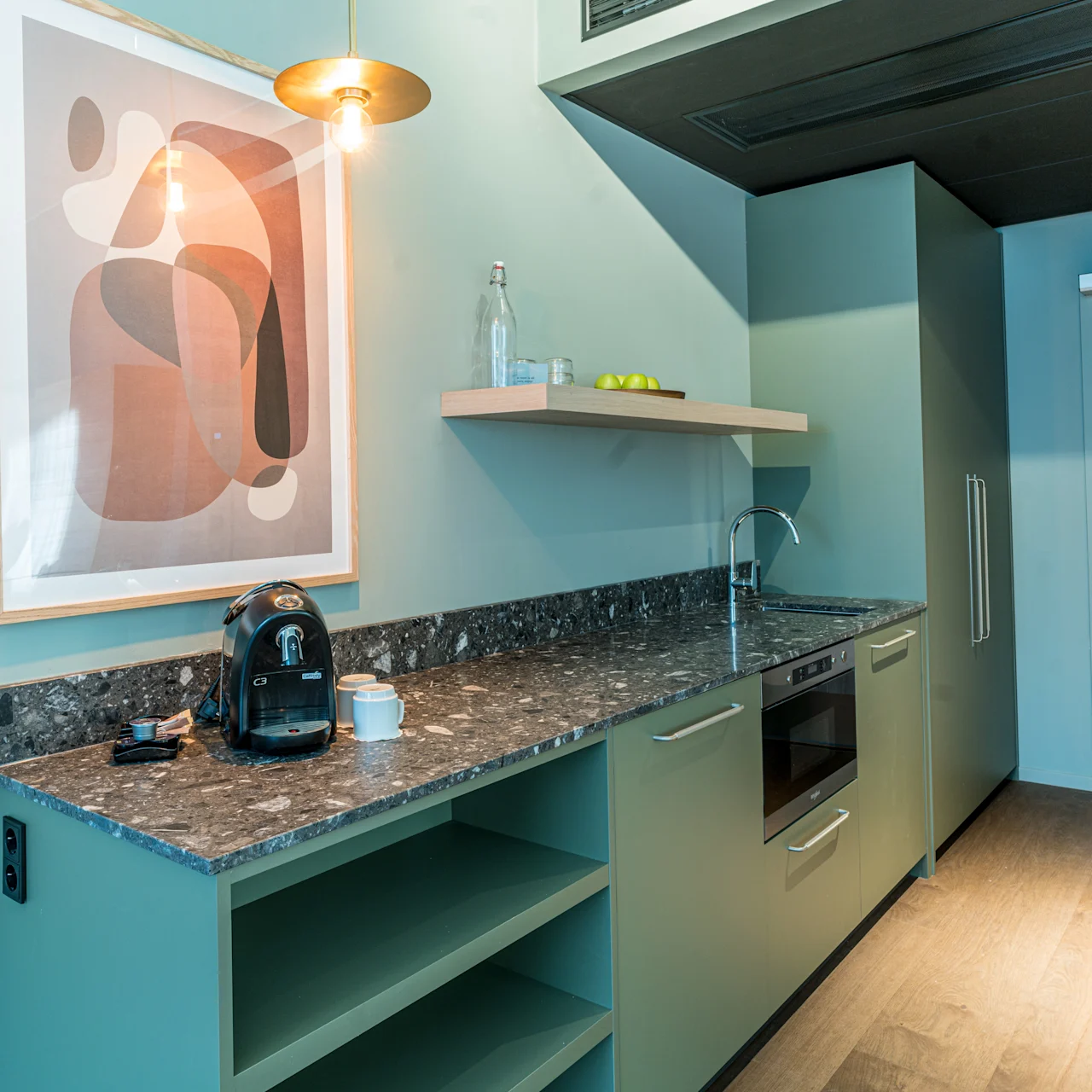 A kitchenette features a coffee machine and cup on a granite countertop. Art hangs on a green wall above. A sink, oven, and shelf with bottles and fruit complete the modern decor.