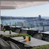 Outdoor terrace with tables and chairs overlooking a scenic waterfront with mountains and city buildings under a clear sky.