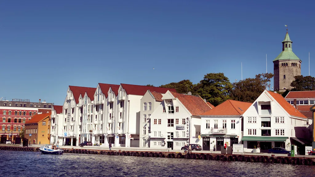 Traditionelle hvide træhuse langs en havnefront under en klar blå himmel med et tårn med grøn top i baggrunden.