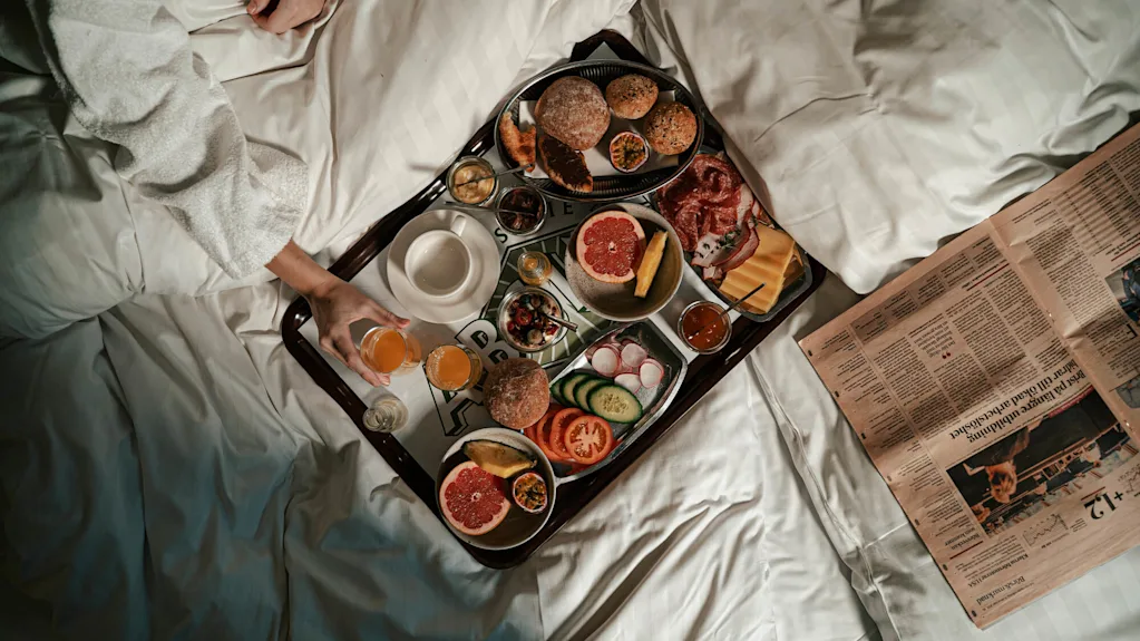 En frukostbricka med bröd, grapefrukt och kött på en säng. En person i morgonrock håller ett glas juice. En tidning ligger bredvid. Text: ”State”.