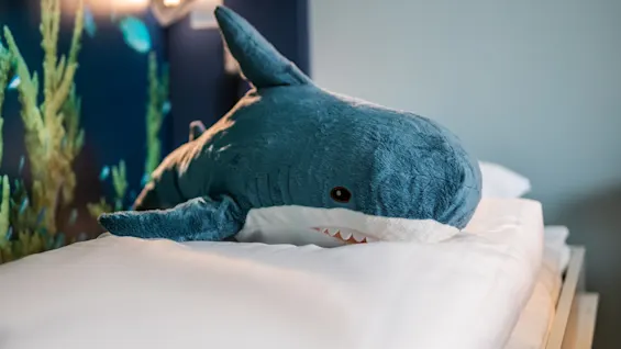 A plush shark rests on a neatly made bed in a room. An aquatic-themed mural is visible on the wall behind, enhancing the underwater theme.