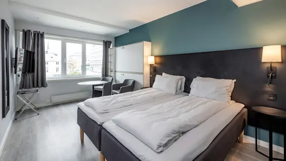 En ordentligt bäddad dubbelsäng med vita sängkläder står bredvid en väggmonterad lampa i ett modernt hotellrum med grå väggar, trägolv och ett fönster med utsikt över staden.