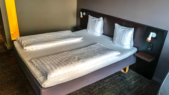En prydligt bäddad hotellsäng med vita randiga sängkläder, kuddar och en mörk sänggavel med integrerad belysning.