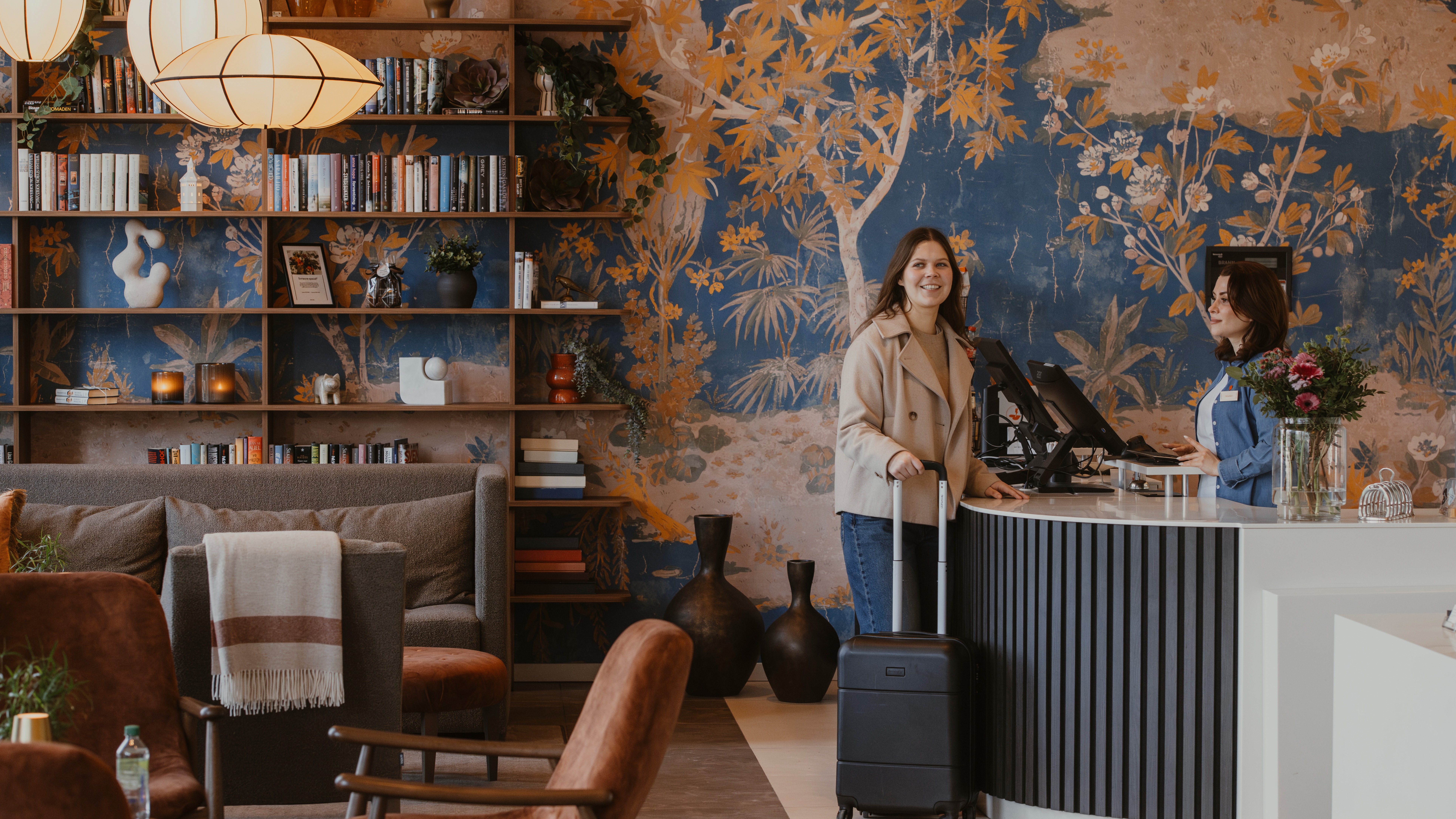 Vieras matkalaukun kanssa hotellin vastaanotossa, vuorovaikutuksessa työntekijän kanssa tyylikkäässä aulassa kirjahyllyn kera.