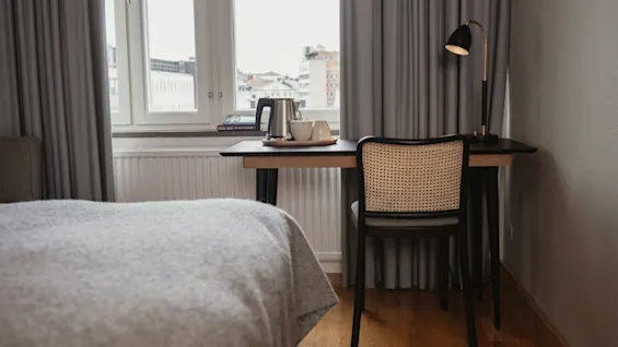 Et skrivebord med en stol holder en lampe, en vannkoker og kopper, med utsikt over et vindu i et svakt opplyst soverom med gardiner og en seng i forgrunnen.