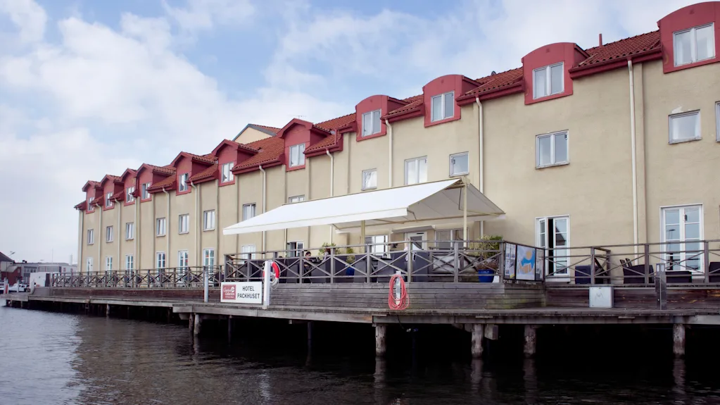 A waterfront building with a terrace extends over water, under a cloudy sky. Sign reads "HOTEL PAKKHUSET".
