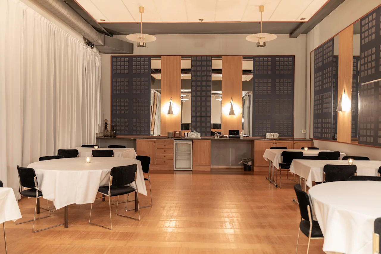 Tilava juhlatila, jossa pyöreät pöydät on katettu vieraille, ja taustalla on tarjoilutiski.