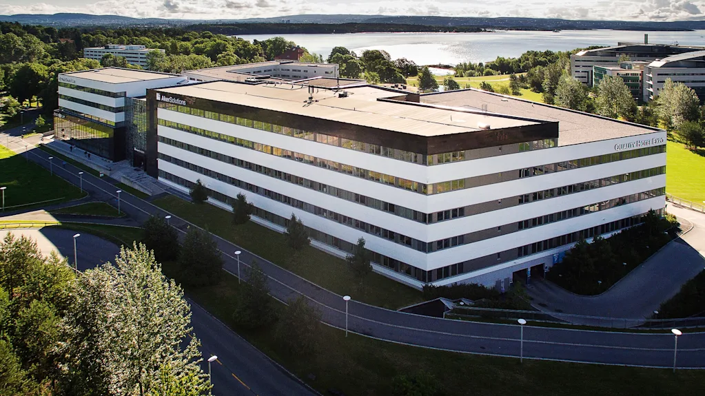 En stor, moderne bygning med "Aker Solutions" på fasaden står blant gressplener og trær, nær et vannlegeme under en klar blå himmel.