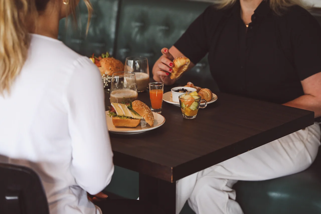 Two people sit at a wooden table enjoying lunch. Sandwiches, coffee, juice, and fruit are served. They are in a casual dining setting with green upholstered seating.
