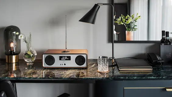 En radio som viser "11:53" og "Matt Spherical - Yuriak" står på et marmor bord med lampe, vase og glass. En telefon og meny merket "IN ROOM SHOP" er i nærheten.