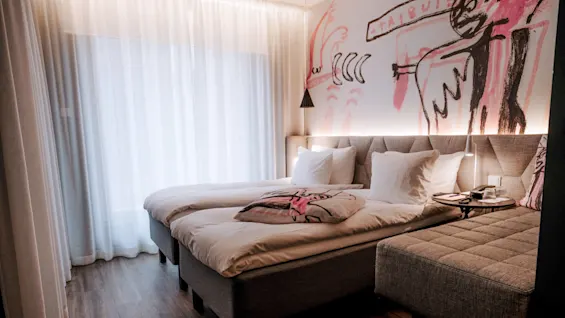 Two neatly made twin beds rest against a wall with abstract pink and black artwork, in a cozy bedroom with sheer curtains and a wooden floor.