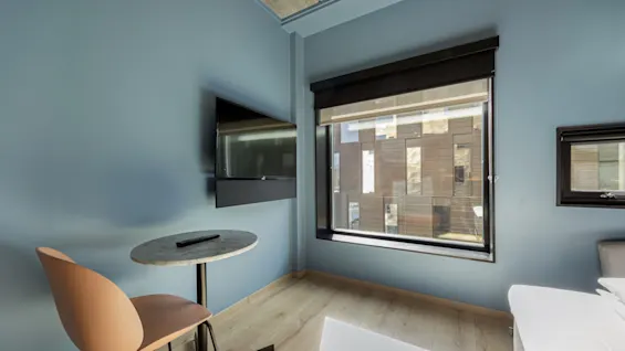 Et lyst, moderne rom med lyseblå vegger, en TV, et lite bord, en stol og vinduer med utsikt.