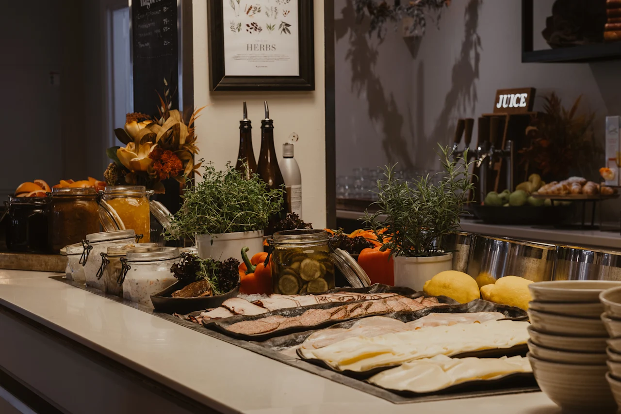 En buffet med forskellige madvarer, herunder pålæg, syltetøj, friske krydderurter og et skilt for HERBS og JUICE.