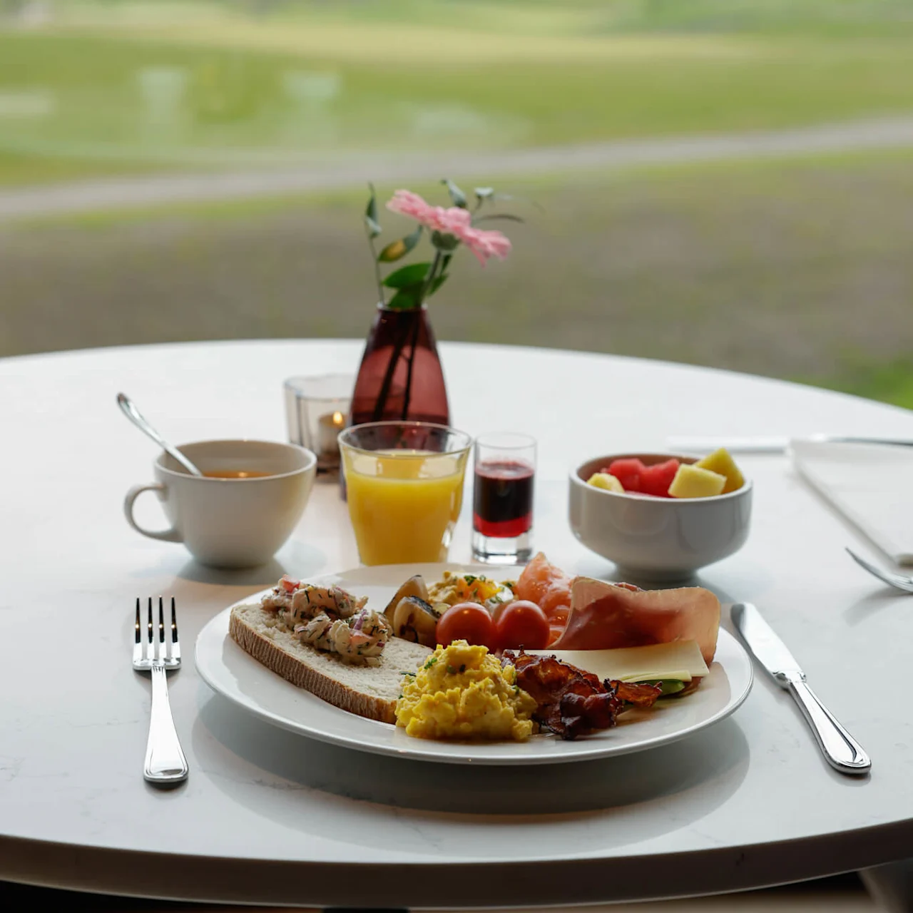 Frukosttallrik med äggröra, kött, och rostat bröd på ett bord, med utsikt över ett grönt landskap på Lily Country Club i norska Klöfta.