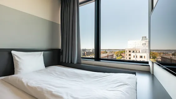 En prydligt bäddad vit säng står bredvid stora fönster, med utsikt över en stadsbild med byggnader under en klar himmel.